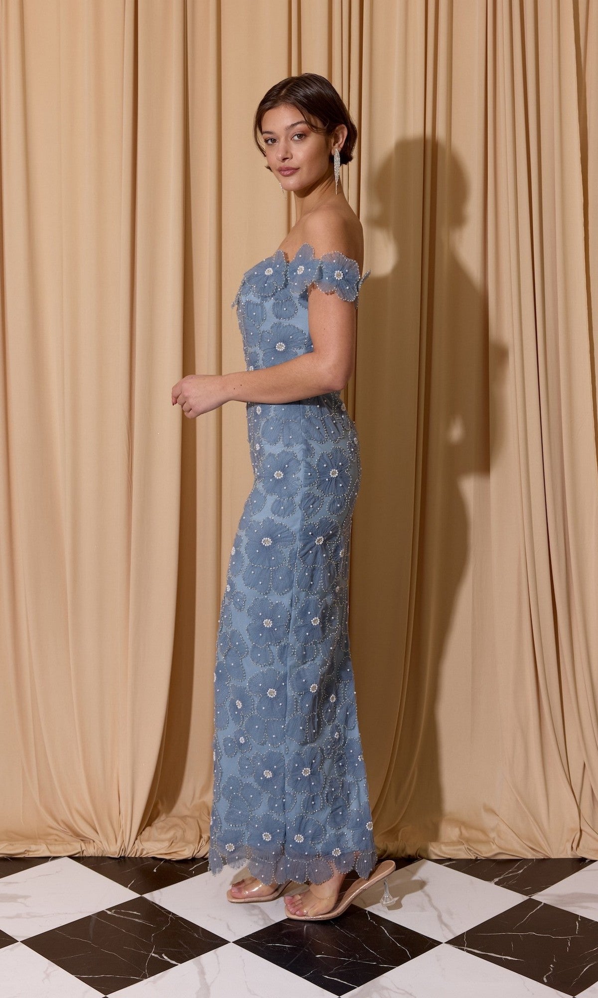 A woman stands sideways in front of beige curtains, modeling the Eureka EK205 Long Prom Dress—an off-the-shoulder blue lace gown with 3D floral patterns—paired with transparent high heels on a black and white tiled floor.